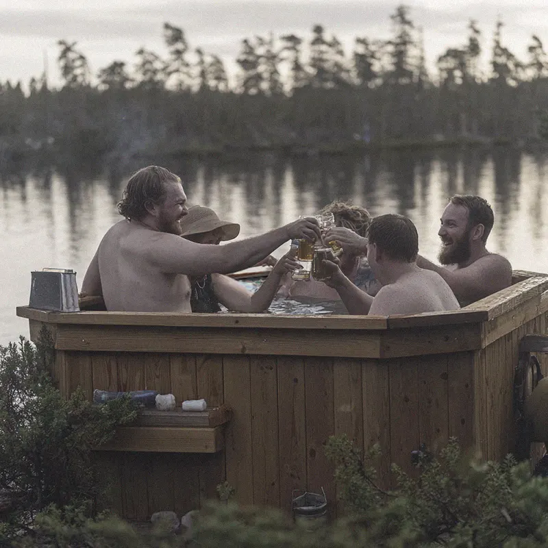 Gruppe genießt entspanntes Bad im Holz-Whirlpool, umgeben von Natur, stoßen mit Getränken an.