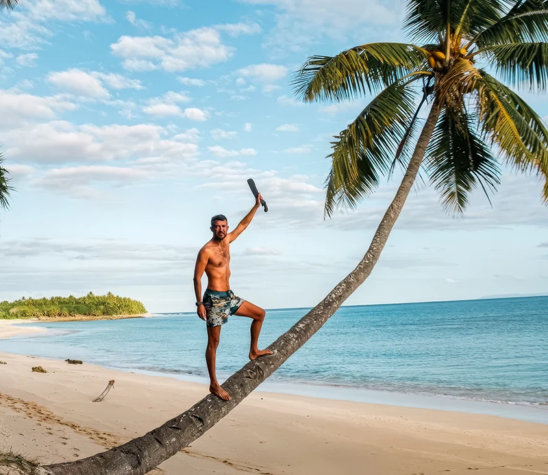 Ein Mann auf einer geneigten Palme am tropischen Strand, hebt einen Arm, blauer Himmel und Meer im Hintergrund.
