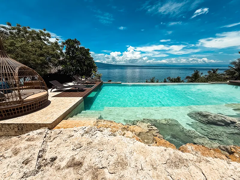 Luxuriöser Infinity-Pool mit Meerblick, umgeben von tropischer Natur und Liegestühlen unter blauem Himmel.