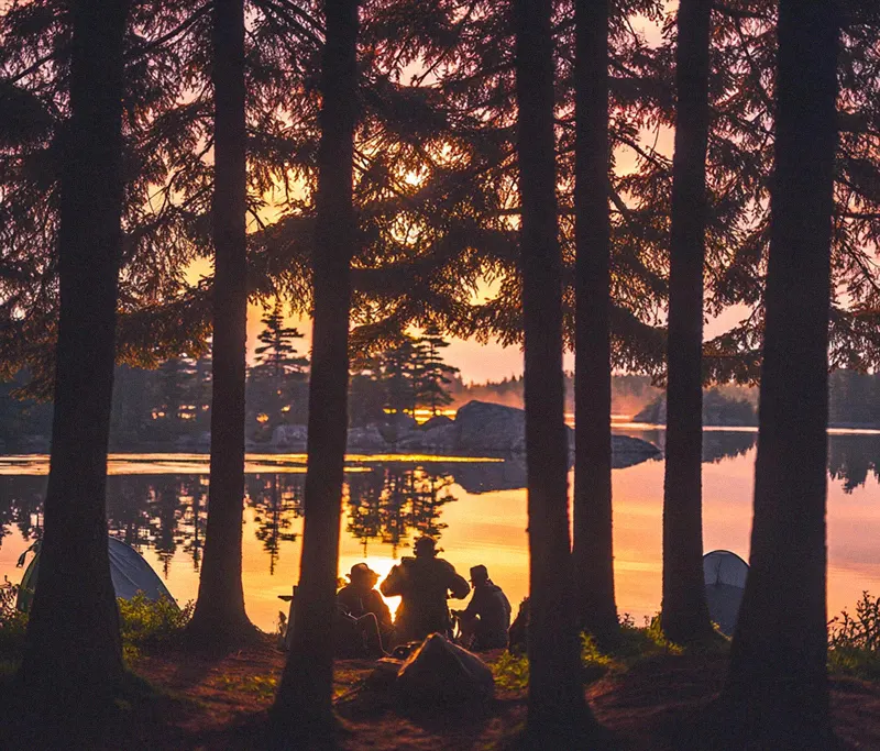 Sonnenuntergang am See hinter Bäumen, Silhouetten von Menschen bei Zelten, friedliche Naturkulisse.