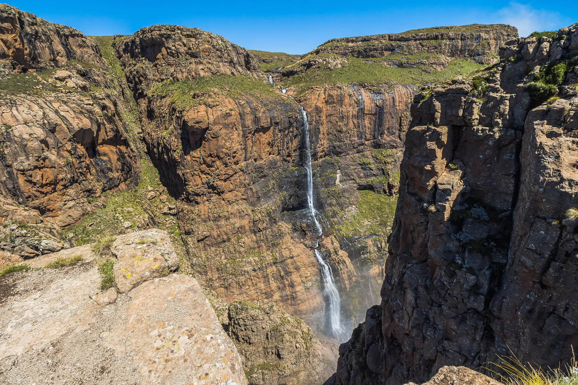 Majestätischer Wasserfall stürzt in tiefen Canyon, umgeben von steilen Felsen und grüner Landschaft unter blauem Himmel.