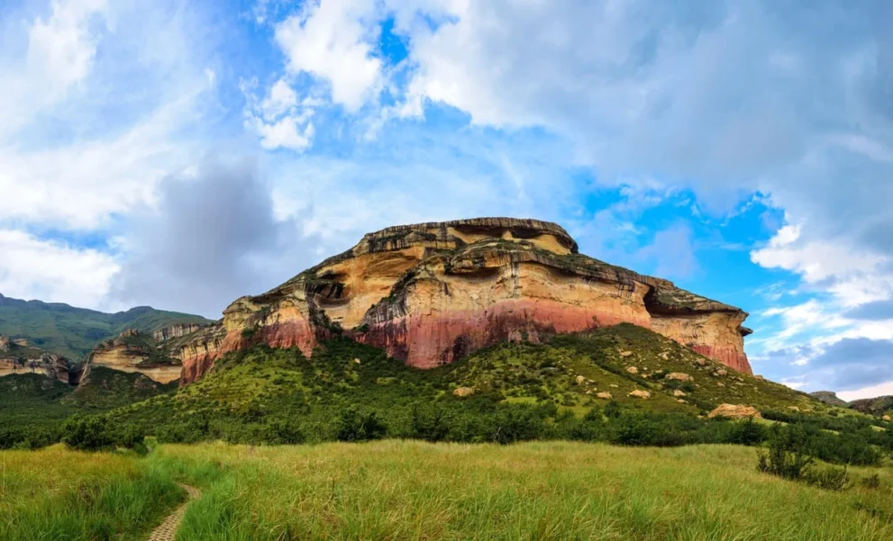 Majestätischer Berg mit bunten Felsen und grünem Vorland unter blauem Himmel voller Wolken. Beeindruckende Naturkulisse.
