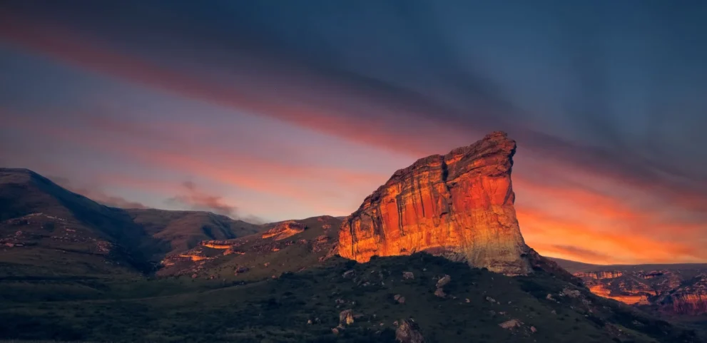 Roter Felsen im Abendlicht vor dramatischem Himmel, umgeben von grünen Hügeln und sanften Landschaften.